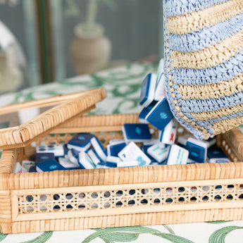 Handwoven rattan Mahjong tile box with glass top, neutral tones, and island-inspired details from the Island Collection. Fits a full-size tile set. With Navy Mahjong tile set from the House Collection, featuring intricately designed tiles with floral motifs, butterflies, and peacocks. Tile size: 1" x 1 7/16" x 7/16".