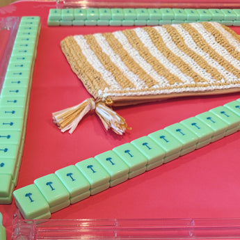 Striped white and tan raffia Mahjong travel bag with tassel, placed on a red game mat surrounded by mint green Mahjong tiles featuring blue palm trees.